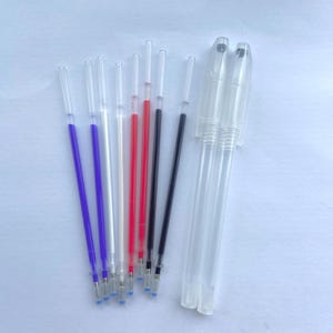 Può includere: Una collezione di ricariche per penne in vari colori, tra cui viola, rosso e nero, accanto a due corpi di penna in plastica trasparente. Le ricariche sono disposte su una superficie bianca, mostrando i loro diversi colori d'inchiostro.