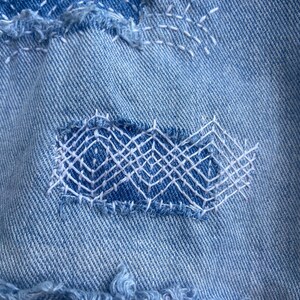 May include: Close-up of light blue denim fabric with visible mending. Patches of denim are sewn onto the base fabric with white thread, creating geometric patterns. The edges of the patches are frayed, adding texture.