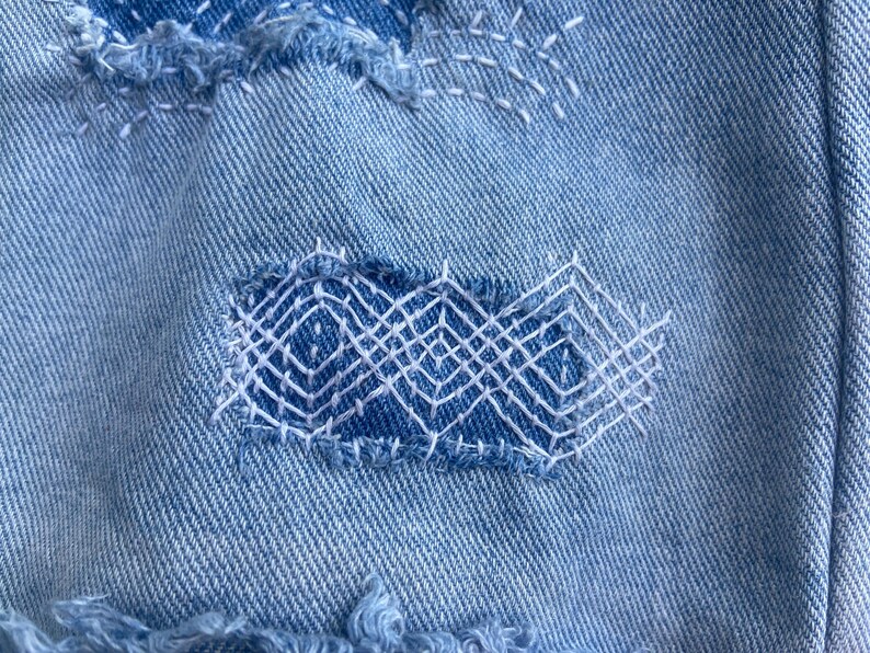 May include: Close-up of light blue denim fabric with visible mending. Patches of denim are sewn onto the base fabric with white thread, creating geometric patterns. The edges of the patches are frayed, adding texture.