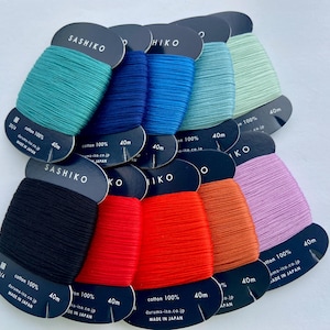 May include: A collection of ten spools of Sashiko thread in various colors including black, red, orange, pink, teal, blue, and light green. Each spool is made of 100% cotton, measures 40m in length, and features the text Sashiko and Made in Japan.