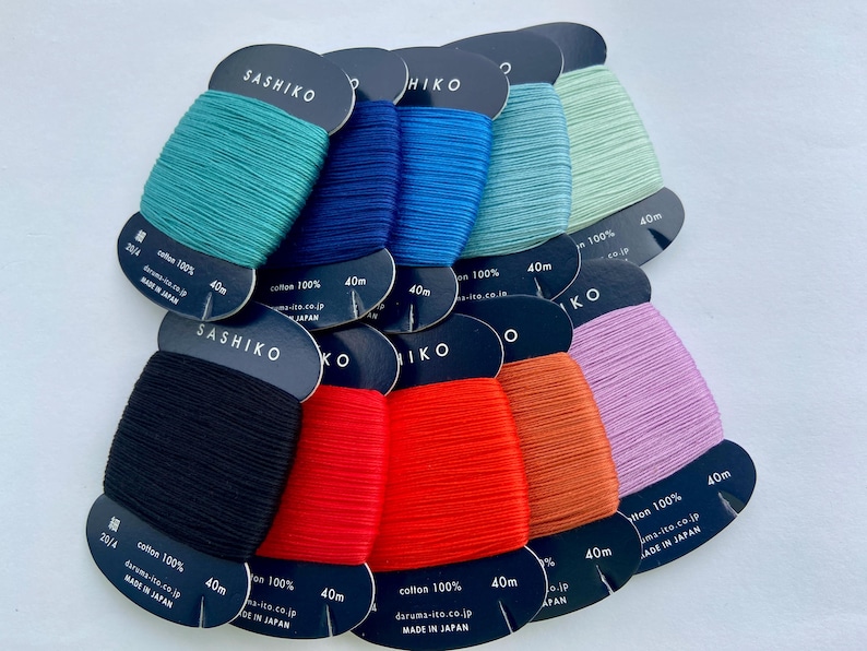 May include: A collection of ten spools of Sashiko thread in various colors including black, red, orange, pink, teal, blue, and light green. Each spool is made of 100% cotton, measures 40m in length, and features the text Sashiko and Made in Japan.