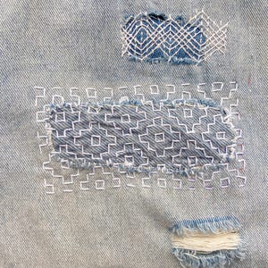 K&ouml;nnte beinhalten: Hellblauer Jeansstoff mit zwei rechteckigen Flicken, die mit wei&szlig;em Garn in einem geometrischen Sashiko-Stickmuster repariert wurden. Ein kleiner, ausgefranster, horizontaler Bereich ist am unteren Rand des Jeansstoffs zu sehen.