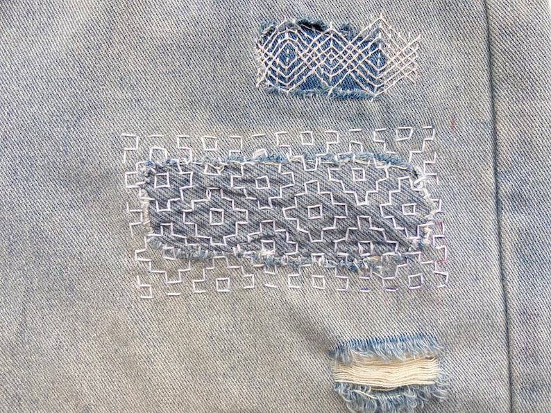 K&ouml;nnte beinhalten: Hellblauer Jeansstoff mit zwei rechteckigen Flicken, die mit wei&szlig;em Garn in einem geometrischen Sashiko-Stickmuster repariert wurden. Ein kleiner, ausgefranster, horizontaler Bereich ist am unteren Rand des Jeansstoffs zu sehen.