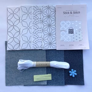 Könnte beinhalten: Ein Bastelset mit Stoffstücken, weißem Faden, einem blauen blütenförmigen Knopf und einem kleinen rechteckigen Stück. Das Set enthält ein Heft mit dem Titel "Stick & Stitch" mit verschiedenen Stichmustern. Die Stoffstücke haben geometrische Designs.