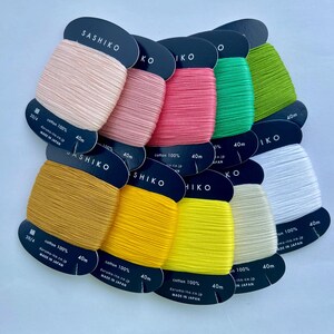 May include: A collection of ten Sashiko embroidery thread spools in various colors including pale pink, rose, hot pink, green, lime, mustard, yellow, bright yellow, cream, and white. Each spool is labeled 100% cotton, 40m, and made in Japan.