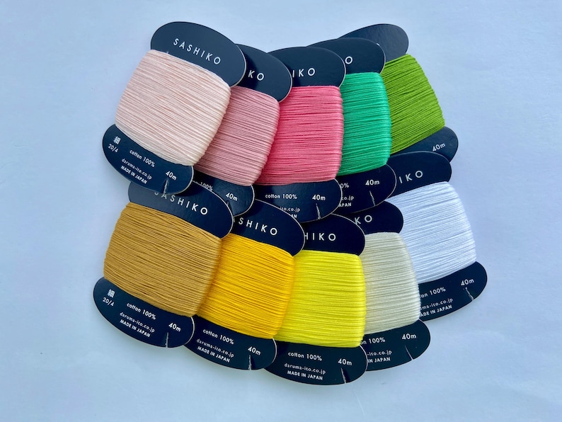 May include: A collection of ten Sashiko embroidery thread spools in various colors including pale pink, rose, hot pink, green, lime, mustard, yellow, bright yellow, cream, and white. Each spool is labeled 100% cotton, 40m, and made in Japan.