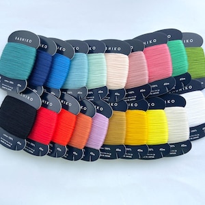 Peut inclure: Une collection de vingt bobines de fil de coton Sashiko, chacune enroulée sur une carte en carton noir indiquant une longueur de 40m. Les fils sont disposés sur deux rangées, présentant un spectre de couleurs vives incluant le bleu sarcelle, le bleu, le rose, le vert, le noir, le rouge, l'orange, le jaune et le blanc.