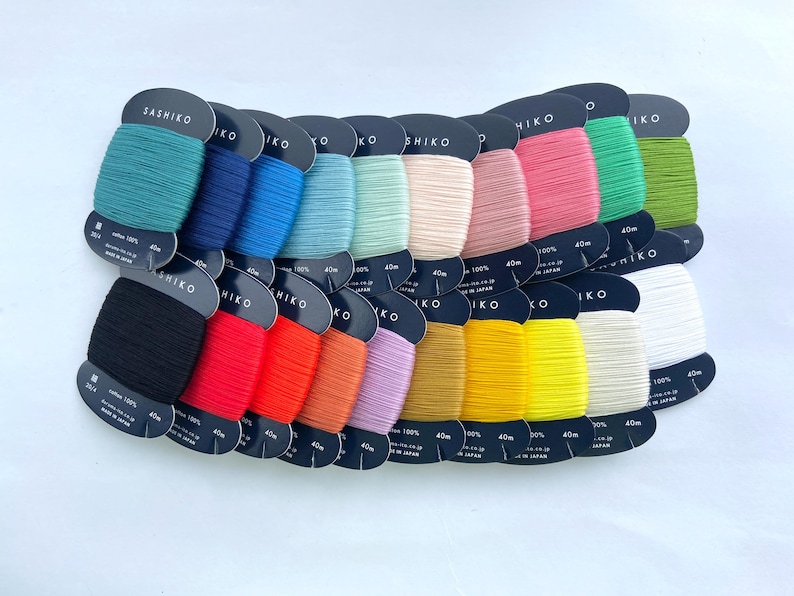 May include: A collection of twenty spools of Sashiko cotton thread, each wrapped on a black cardboard card labeled with 40m length. The threads are arranged in two rows, displaying a vibrant spectrum of colors including teal, blue, pink, green, black, red, orange, yellow, and white.