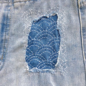 K&ouml;nnte beinhalten: Hellblaue Jeansshorts mit einer abgenutzten rechteckigen Stelle, die mit dunkelblauem Stoff repariert wurde. Der Flicken ist mit wei&szlig;en Sashiko-N&auml;hten in einem sich wiederholenden F&auml;chermuster verziert. Die Kanten des Jeansstoffs sind ausgefranst.