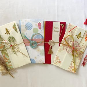May include: Four decorative Japanese envelopes with intricate designs and embellishments. The envelopes feature various colors, including cream, red, and blue, with gold accents and decorative knots. One envelope has the text "Happy Wedding".