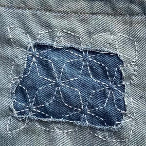 May include: A light blue denim fabric featuring a rectangular dark blue patch sewn onto the surface. The patch is decorated with white hand-stitched geometric patterns. The top edge of the denim shows a frayed, distressed hemline detail.