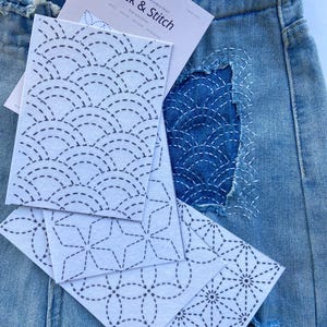 May include: A pair of light blue denim jeans with a frayed waistband and a distressed patch featuring white stitched geometric patterns. Several white water-soluble embroidery sheets labelled Stick & Stitch with black printed designs are layered on top.