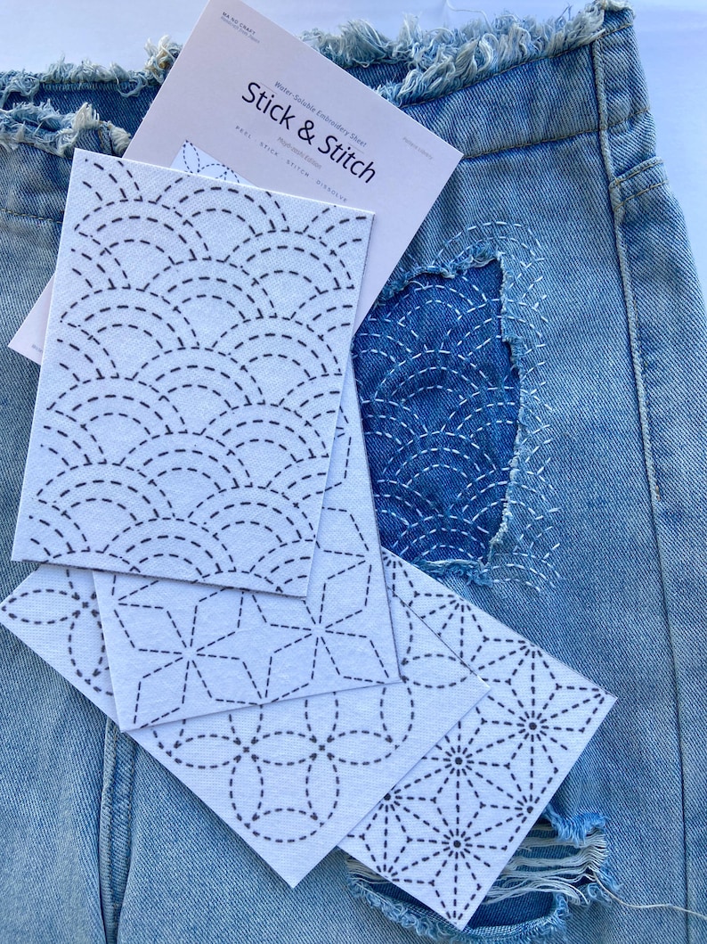 May include: A pair of light blue denim jeans with a frayed waistband and a distressed patch featuring white stitched geometric patterns. Several white water-soluble embroidery sheets labelled Stick & Stitch with black printed designs are layered on top.