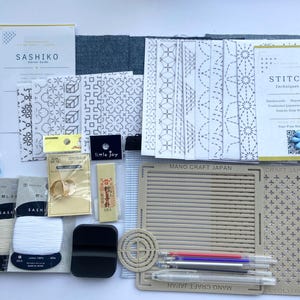 Puede incluir: Un kit de bordado Sashiko que incluye hilo blanco, telas con patrones impresos, un estuche para agujas, dedales, una herramienta de marcado amarilla, una herramienta negra, una rejilla de plástico beige, una guía circular y bolígrafos con recambios.