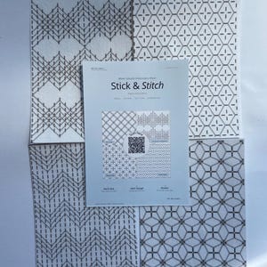 May include: Several white fabric squares featuring grey stitched designs. A central sheet of paper is labelled "Stick & Stitch", detailing the product's use. The designs include geometric and chevron patterns, suitable for embroidery projects.