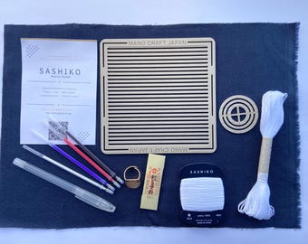 Puede incluir: Un kit de inicio para bordado Sashiko que incluye un cuadrado de tela azul oscuro, una plantilla rectangular con ranuras horizontales, una plantilla circular, hilo blanco, un dedal, agujas y cuatro rotuladores de colores con una guía.