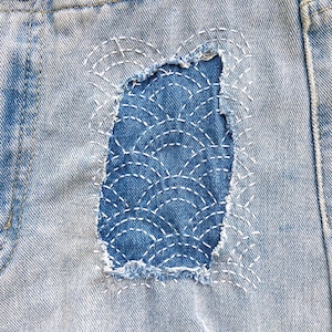 May include: Light blue denim shorts featuring a distressed rectangular patch area repaired with dark blue fabric. The patch is decorated with white sashiko-style stitching in a repeating fan pattern. The edges of the denim are frayed and worn.