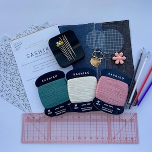 Puede incluir: Un kit de inicio de Sashiko con varios suministros. Incluye carretes de hilo verde, blanco y rosa, agujas, muestras de tela, una regla y bolígrafos. El kit también contiene una guía con la palabra "Sashiko" en la portada.