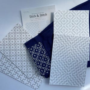 Puede incluir: Un conjunto de cuadrados de tela blanca con patrones geométricos, junto con una tela azul marino y una hoja "Stick & Stitch". Los diseños incluyen motivos de cruces y cuadrados, para proyectos de bordado.