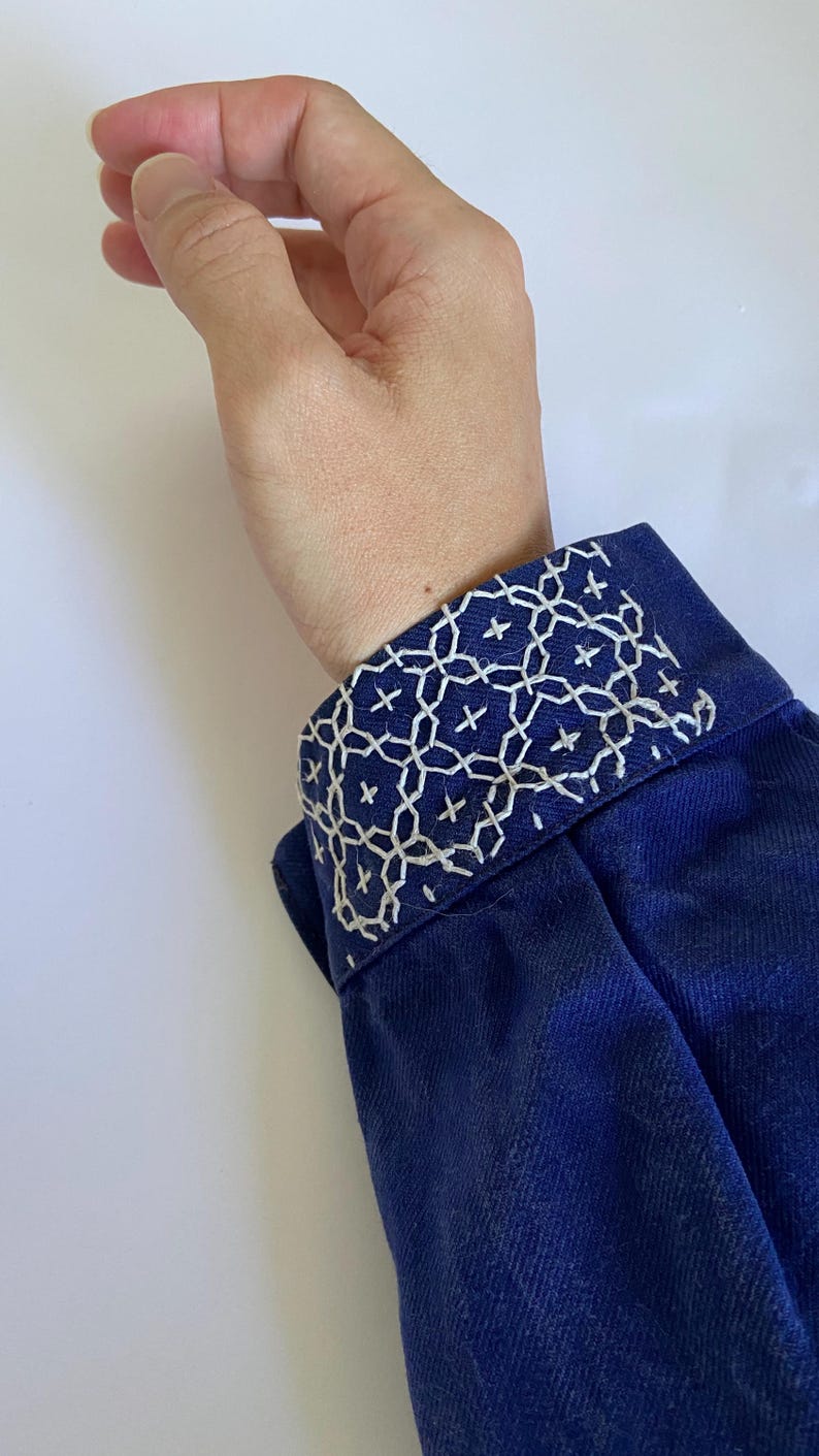 K&ouml;nnte beinhalten: Ein dunkelblaues lang&auml;rmeliges Kleidungsst&uuml;ck mit einer strukturierten Manschette, die mit aufwendiger wei&szlig;er geometrischer Stickerei verziert ist. Die Stiche bilden ein wiederkehrendes Muster aus ineinandergreifenden Kreisen und kleinen Kreuzen.