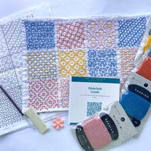 Può includere: Un kit per ricamo Sashiko con un campione di tessuto bianco con motivi geometrici blu, gialli e rosa, una guida Dishcloth con codice QR, quattro rocchetti di filo, un ago sottile in metallo, un gesso rettangolare e una perlina a forma di fiore.