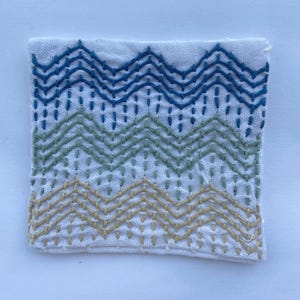 May include: A square of white fabric with hand-stitched embroidery. The embroidery features a chevron pattern in shades of blue, green, and tan. The stitches are evenly spaced, creating a textured design. The fabric has a slightly wrinkled appearance.