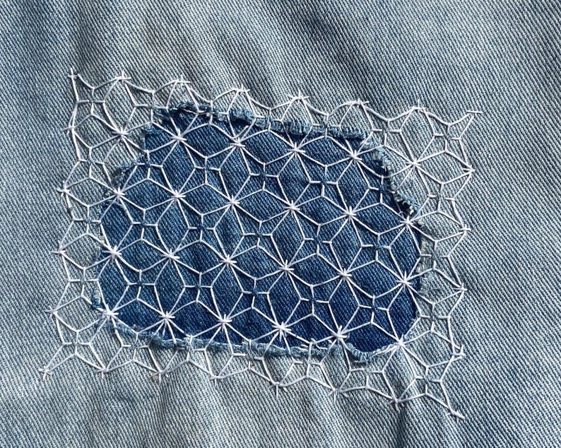 May include: Close-up of a denim patch with intricate white embroidery. The patch features a geometric design over a blue fabric, stitched onto a light blue denim background. The embroidery adds a decorative element to the denim.