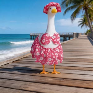 May include: A white goose figurine wearing a pink floral print bikini and matching sun hat. The goose stands on a wooden boardwalk with a beach and palm trees in the background. The goose has orange feet and a yellow beak.