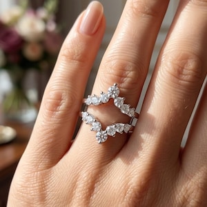 Peut inclure: Une bague en argent avec un design en forme de V, ornée de plusieurs petites pierres précieuses rondes et transparentes. La bague est portée sur un doigt, mettant en valeur ses détails étincelants. L'arrière-plan est flou.