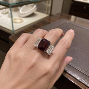 May include: A three-stone ring featuring a large, rectangular, deep red gemstone in the center, flanked by two smaller, rectangular, clear white gemstones on either side, all set in a polished silver-toned metal band.