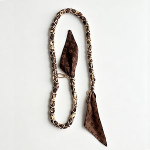 May include: A long, flexible strap featuring a gold-tone metal chain intertwined with a brown fabric ribbon. The ribbon displays a repeating pattern of small flowers and geometric shapes. Two triangular fabric ends hang from gold-tone metal clasps.