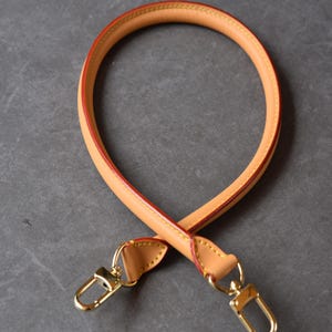 May include: Tan leather handbag handle with gold-tone metal clasps. The handle is round with a red edge and yellow stitching. The clasps are attached to the handle with matching tan leather.