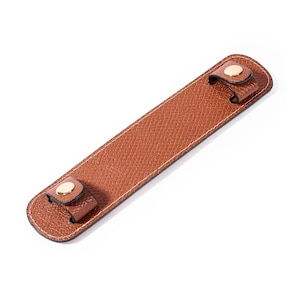 May include: A rectangular brown leather handle wrap with a textured surface and white stitching along the edges. The wrap features two small loops at each end, each secured with a polished gold-tone metal snap button fastener for closure.