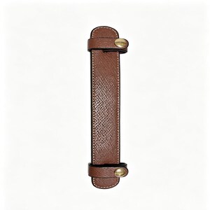 May include: A rectangular brown leather handle wrap with a textured surface and white stitching along the edges. The wrap features two smaller leather loops at the top and bottom, each secured with a circular gold-toned metal snap button fastener.