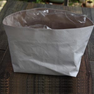 May include: A light gray, open-top tote bag with a textured surface. The interior is a lighter beige color, and it contains clear plastic packaging. The bag is sitting on a wooden surface.