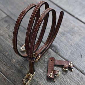 May include: A brown leather purse strap with gold-colored hardware. The strap is stitched with a contrasting thread and has a swivel clasp. A separate leather piece with gold hardware is also included.