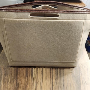 Puede incluir: Organizador de bolso de fieltro beige con un bolsillo rectangular en la parte delantera. El borde superior tiene dos asas y está ribeteado con cuero marrón. El organizador está diseñado para caber dentro de un bolso más grande.