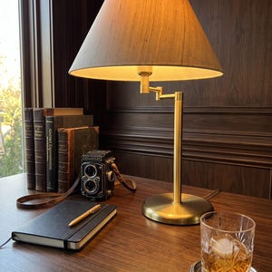 Könnte beinhalten: Eine Schreibtischlampe aus Messing mit konischem Stoffschirm, eine alte Rolleiflex-Zweiäugige Spiegelreflexkamera, ein schwarzes Notizbuch mit goldenem Stift und ein Kristallglas mit bernsteinfarbener Flüssigkeit und Eis.