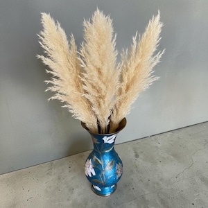 May include: A blue metal vase with a flared rim and a rounded body, decorated with white and gold floral patterns. The vase contains several tall, fluffy, beige pampas grass plumes that extend upwards in a fan-like arrangement from the opening.