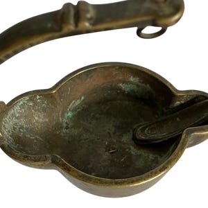 Peut inclure: Une lampe à huile en laiton terni dotée d'un bol en forme de trèfle avec une oxydation verte visible et une poignée incurvée s'étendant sur le côté. La surface métallique montre des signes d'âge et d'usure, avec une petite languette à l'intérieur.