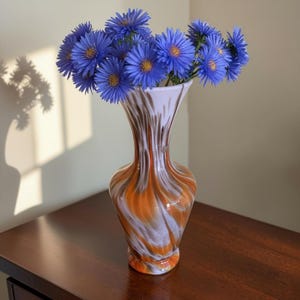 Può includere: Un vaso in vetro con bordo svasato e collo affusolato, caratterizzato da un motivo marmorizzato a vortice nei toni del bianco, dell'arancione e del marrone. Il vaso contiene un bouquet di vivaci fiori blu con centri gialli.