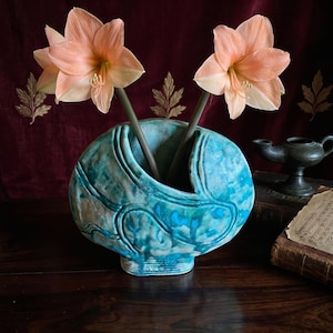 Op de afbeelding: Een turquoise en blauwe keramische vaas met een afgeronde, organische vorm en textuurdetails op het oppervlak. Twee perzikkleurige amaryllisbloemen met lange groene stelen staan in de vaas en steken uit de centrale opening omhoog.
