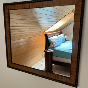 May include: A rectangular wall mirror featuring a wide, light brown wooden frame with a dark, textured outer edge. The mirror surface reflects a bedroom interior with a wooden bed frame, light blue bedding, and a small bedside table with a lamp.