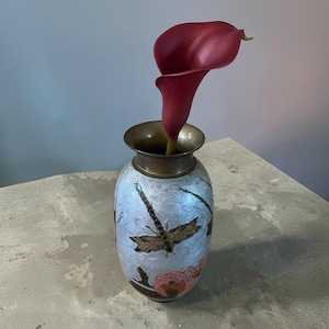 Op de afbeelding: Een kleine, ronde metalen vaas met een lichtblauwe, getextureerde afwerking met een donkere libel en roze bloemmotieven. De vaas heeft een metalen rand en bevat een enkele, dieprode calla-lelie die uit de smalle opening aan de bovenkant steekt.