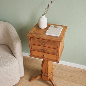 Peut inclure: Une table d'appoint en bois dotée de trois tiroirs, chacun muni d'une petite poignée en laiton. La table possède un plateau carré soutenu par un pied central à trois branches. Un vase blanc avec une tige séchée et un livre intitulé DESIGN MONOGRAPHS sont posés dessus.
