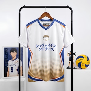May include: A white V-neck sports jersey featuring blue and gold accents, the text "ADLERS" with an eagle logo, and Japanese characters "シュヴァイデン アドラーズ" printed on the front. The design includes a gradient dot pattern at the bottom and blue side panels.