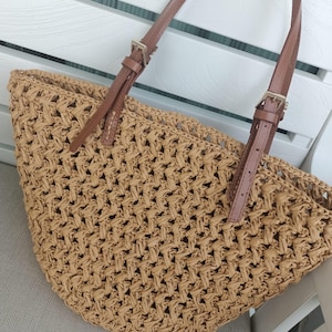 May include: A woven straw tote bag with brown leather straps and silver-tone buckles. The bag has a semi-circular shape and a textured, open weave design. The straps are attached to the bag with metal hardware.