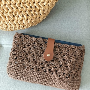 Op de afbeelding: Een bruine gehaakte clutch met een bloemenpatroon en een leren bandsluiting. De clutch is gemaakt van een zacht, geweven materiaal. Op de achtergrond een bijpassende geweven tas.