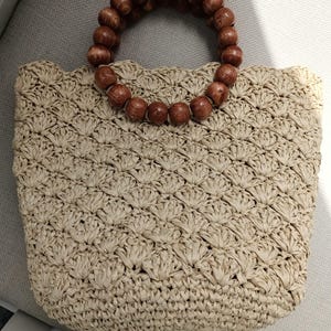 May include: A beige woven handbag with a scalloped top edge. The bag features a circular handle made of dark brown wooden beads. The woven pattern creates a textured surface, and the bag is sitting on a light gray surface.