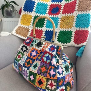 May include: A colorful, handmade crochet bag with a bamboo handle rests on a gray armchair. The bag features a patchwork design of various colors, including blue, green, red, and orange. A matching crochet blanket with a similar pattern is in the background.